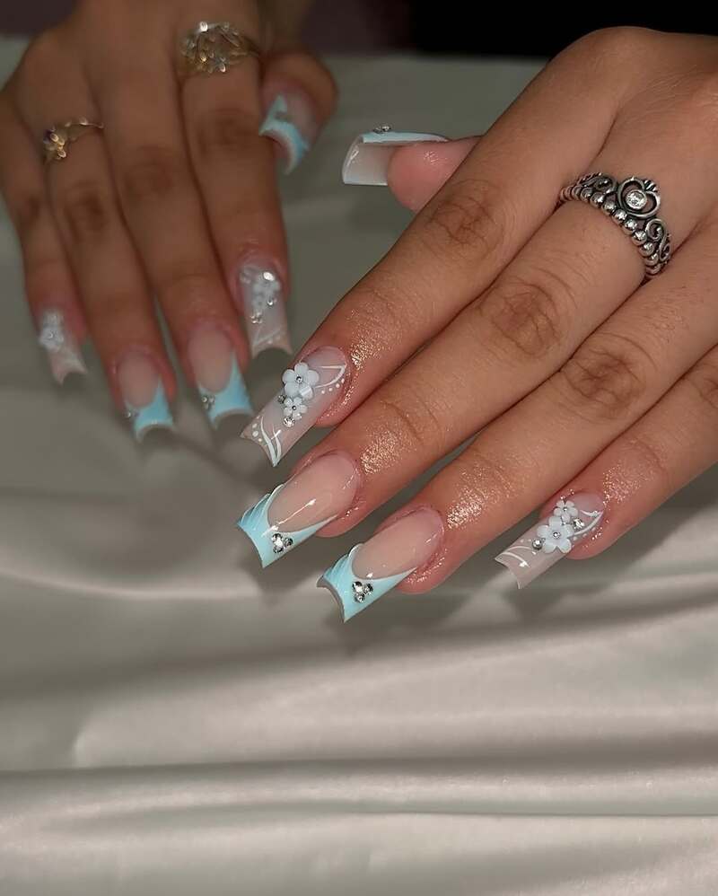 Light blue French tip acrylic nails with white floral designs and rhinestones