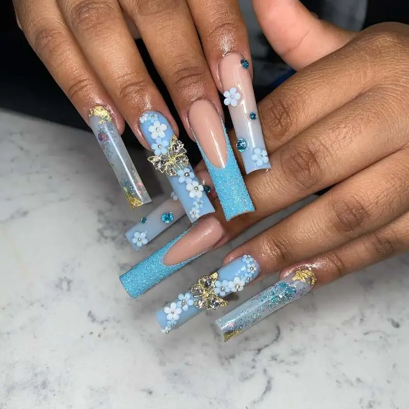 Long nails featuring blue and white floral designs, butterfly charms, glitter accents, and crystal embellishments