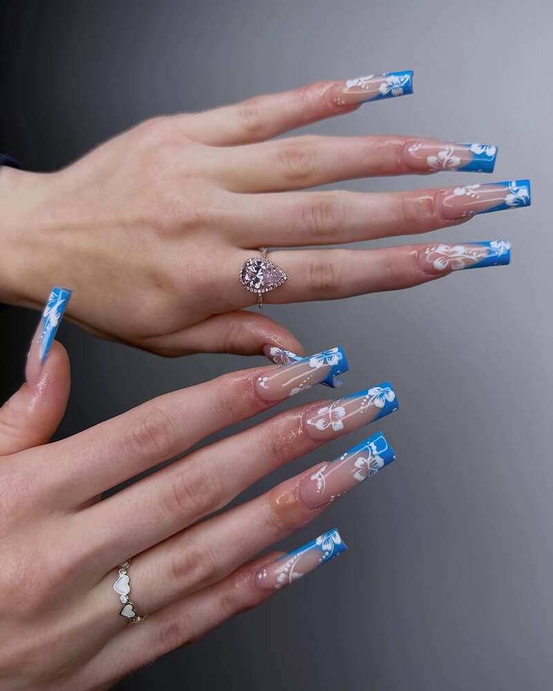 Long square nails with blue French tips and white hibiscus flower designs on clear base